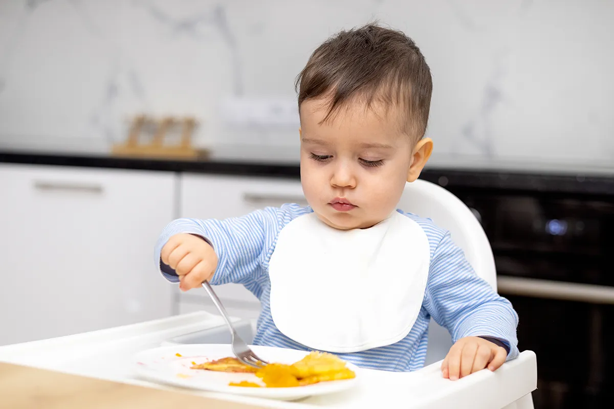 Jeune enfant mangeant seul avec une fourchette dans sa chaise haute.